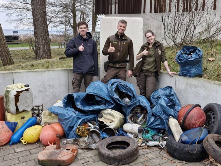 Drei Personen stehen hinter einem Haufen aus Müll und reckten jeweils einen Daumen vor der Brust nach oben.